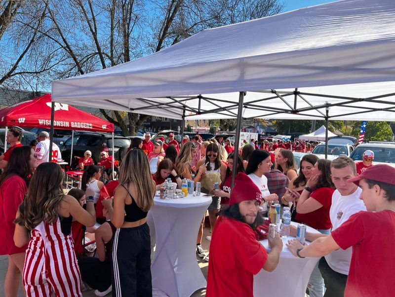 Packed fan zone of Badgers fans around cocktail tables under GameDay Your Way tents on Regent Street.